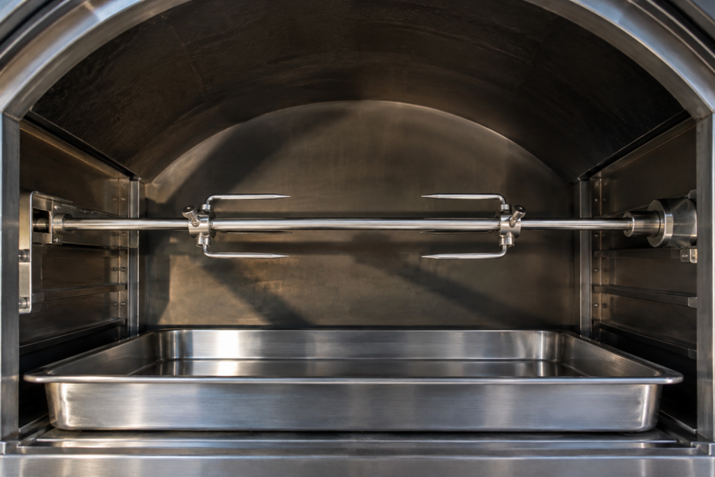 The inside of a cold vaulted oven cavity with a steel rotisserie spit mounted across it between two pronged forks and a drip tray below.