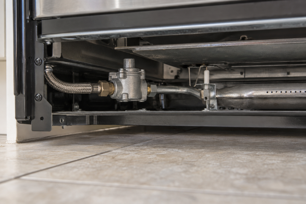 The gas inlet end of an oven burner tube with a small valve body and a braided connector, lower panel removed.