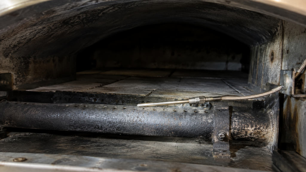 A thin thermocouple probe and lead wire lying across a cast-iron burner tube inside a cooled arched oven.